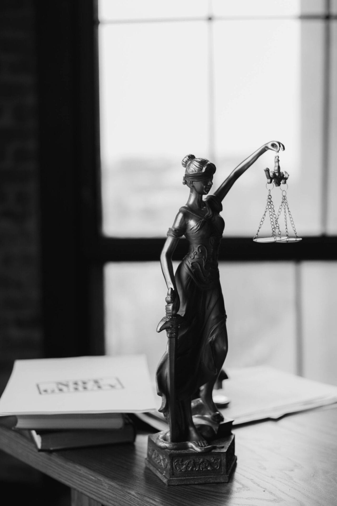 Grayscale photo of Lady Justice statue beside legal documents on a wooden table.
