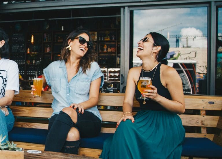 Two women laughing and enjoying drinks outdoors at a trendy bar in Baltimore, creating a vibrant and social atmosphere.