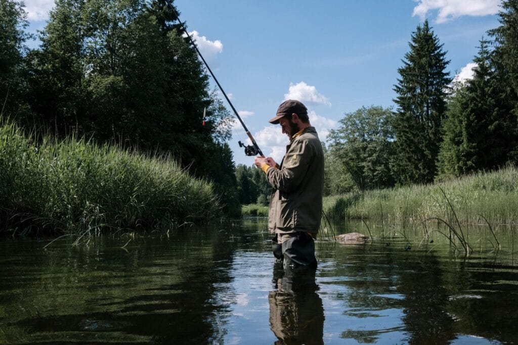 A man in outdoor gear enjoys fishing in a serene forest river under a clear blue sky.